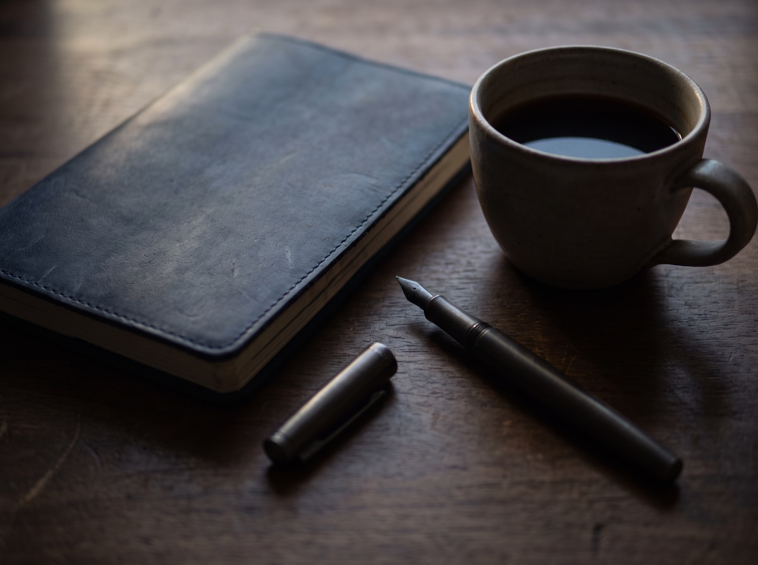 A worn leather notebook, an uncapped fountain pen and a cup of coffee on a wooden desk